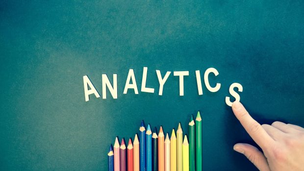 The word analytics against a teal background. There are colored pencils under the word